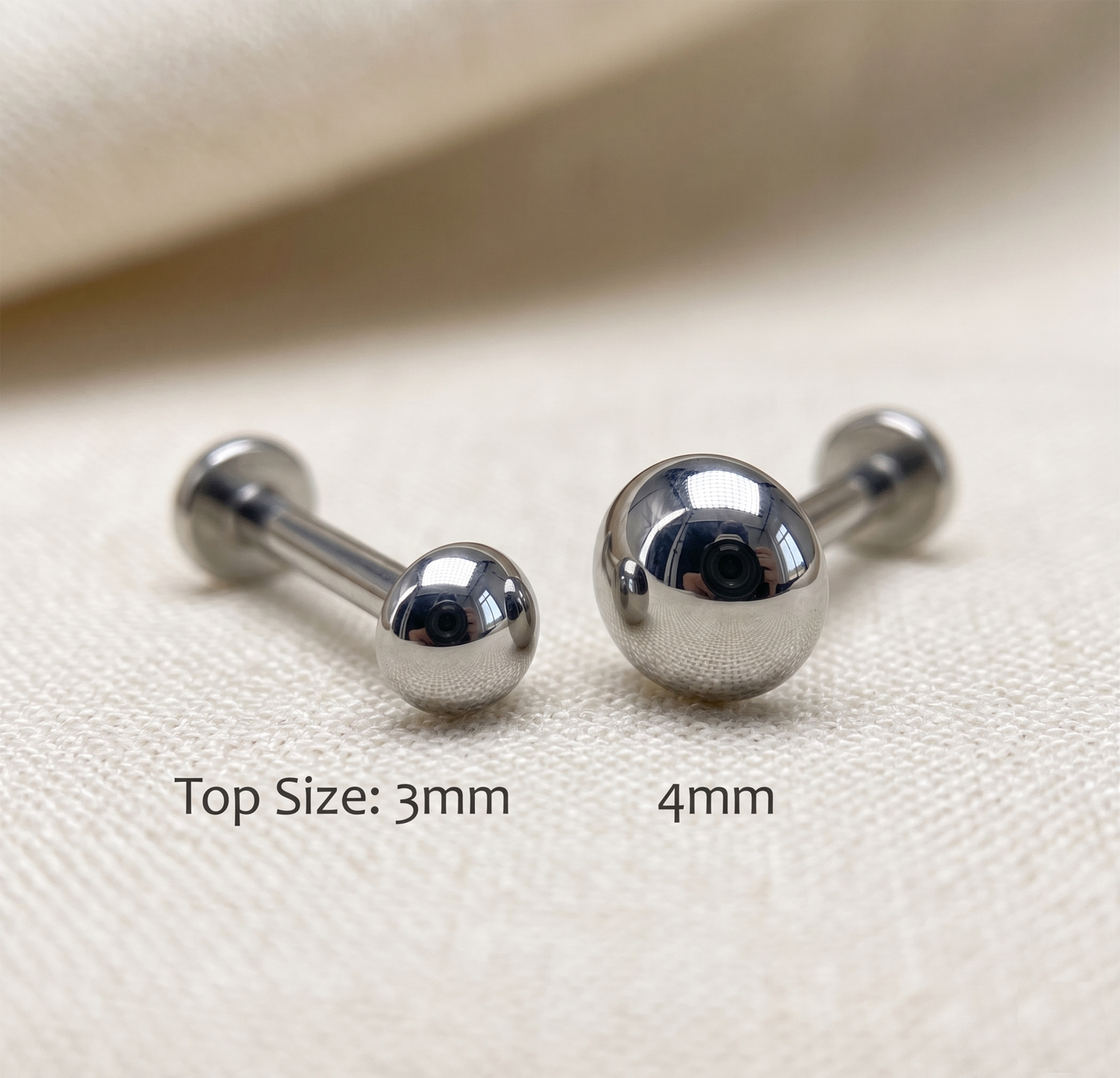 Two silver barbell earrings on a beige fabric background with size labels.