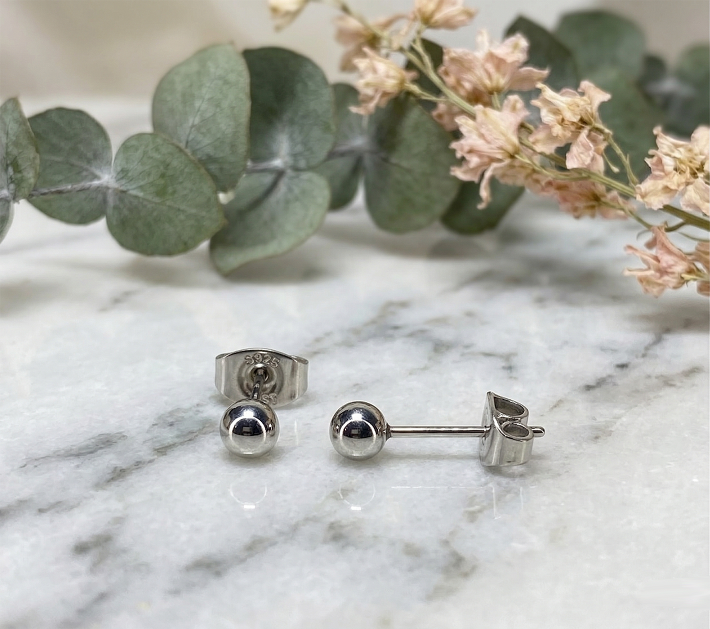 Silver surgical steel ball stud earrings on marble surface with greenery