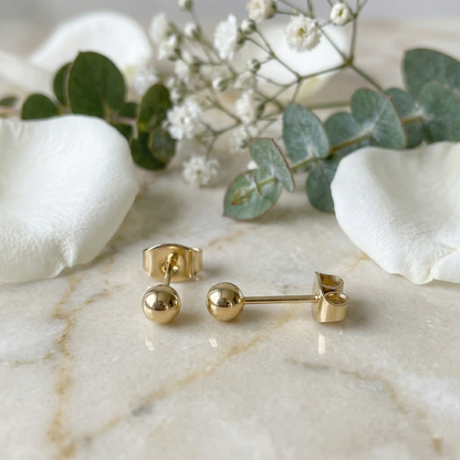 Gold titanium ball stud earrings on white surface with flowers