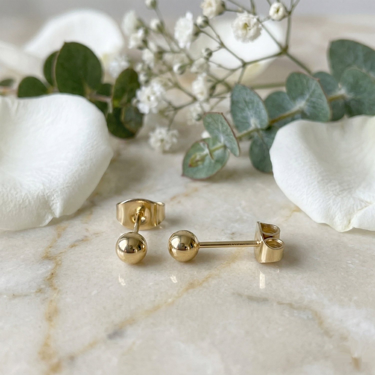 Gold surgical steel ball stud earrings on white surface with flowers
